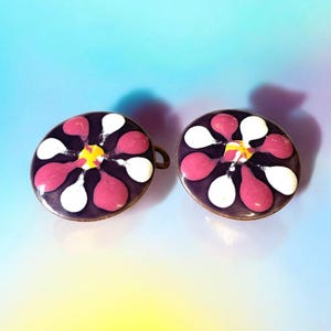 Può includere: Un paio di orecchini rotondi a forma di fiore in smalto viola, rosa, bianco e giallo. Gli orecchini hanno un design semplice.