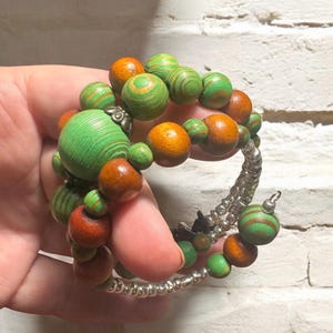 May include: A spiral bracelet with wooden beads in shades of green and orange. The beads are various sizes and shapes, some with striped patterns. The bracelet is held together with a silver-colored wire.