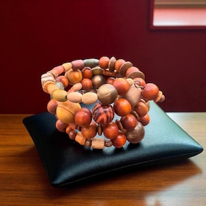 May include: A multi-strand bracelet with a variety of wooden beads in shades of brown, orange, and tan. The beads are different shapes and sizes, including round, oval, and cylindrical. The bracelet is displayed on a black square pillow.