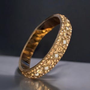 Può includere: Un braccialetto rigido dorato con una fila di piccoli strass trasparenti e strass bianchi più grandi.