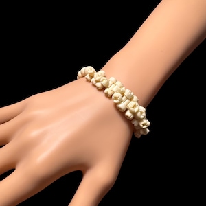 Könnte beinhalten: Elfenbeinfarbenes Armband mit floralem Design. Das Armband besteht aus kleinen, blütenförmigen Perlen, die aneinandergereiht sind. Es wird auf einer Mannequin-Hand vor schwarzem Hintergrund gezeigt.