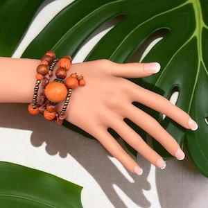 May include: A multi-strand bracelet featuring wooden beads in shades of orange and brown. The bracelet includes various sizes and shapes of beads, with a central large, round bead. The bracelet is displayed on a mannequin hand.