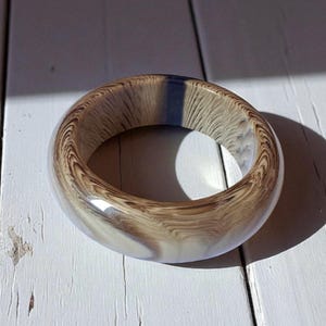 Puede incluir: Un brazalete ancho y redondo con un patrón de vetas de madera en tonos marrón y crema. El brazalete tiene un acabado brillante y está colocado sobre una superficie de madera blanca. El diseño recuerda a la madera natural.