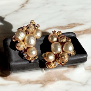 May include: A pair of gold tone earrings with faux pearls and rhinestones. The earrings are shaped like a cluster of pearls.