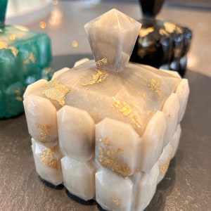 May include: A small, square, grey and white box with a geometric design. The box has a lid with a pointed top and gold accents. The box is decorated with gold leaf.