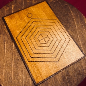 Könnte beinhalten: Ein Holzuntersetzer mit geometrischem Design. Der Untersetzer hat eine hellbraune, rechteckige Mitte mit einer Reihe konzentrischer, schwarzer sechseckiger Formen. Die Mitte befindet sich auf einer größeren, runden, dunkelbraunen Holzbasis.