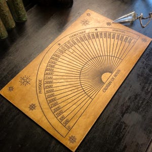 Könnte beinhalten: Ein hölzernes Wünschelrutenbrett mit fächerförmigem Design und schwarzen geätzten Markierungen. Das Brett zeigt die Wörter "Physique", "Energetique" und "Spirituel" sowie numerische Skalen und den Text "Cadran de Bovis". Ein Kristallpendel hängt an dem Brett.