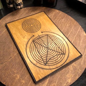 Könnte beinhalten: Ein Holzuntersetzer mit geometrischem Design. Der Untersetzer hat eine hellbraune Oberfläche mit schwarzem Rand. Das Design zeigt konzentrische Kreise und ein sternförmiges Muster. Der Untersetzer liegt auf einer größeren, runden Holzoberfläche.