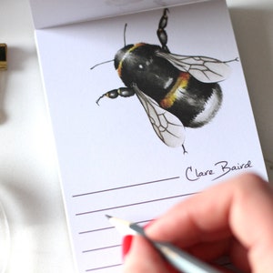 Op de afbeelding: Een wit notitieboekje met een illustratie van een zwart-gele hommel. Het notitieboekje heeft gelinieerde pagina's en de tekst "Clare Baird" staat onderaan. Een hand houdt een potlood vast en schrijft in het notitieboekje.
