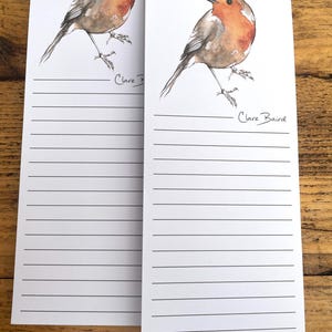May include: Two white notepads with horizontal lines and a watercolor illustration of a robin. The bird has a red breast, brown wings, and is perched on a thin branch. The artist's signature, "Clare Baird," is below the bird.