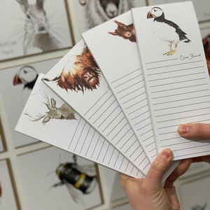 May include: A set of five white notepad memo pads with lined paper. Each pad features a different animal illustration in watercolor style: a puffin, a highland cow, a deer, a squirrel, and a rabbit. The pads are fanned out in a hand, with the artist's signature "Clare Baird" visible on the puffin pad.