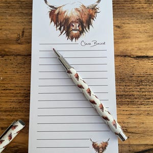 Pu&ograve; includere: Un blocco note bianco con un'illustrazione di mucca Highland marrone e bianca in alto. Il blocco note ha carta rigata e una penna argentata con un motivo di mucca Highland marrone e bianca. Il blocco note &egrave; firmato "Clare Baird".