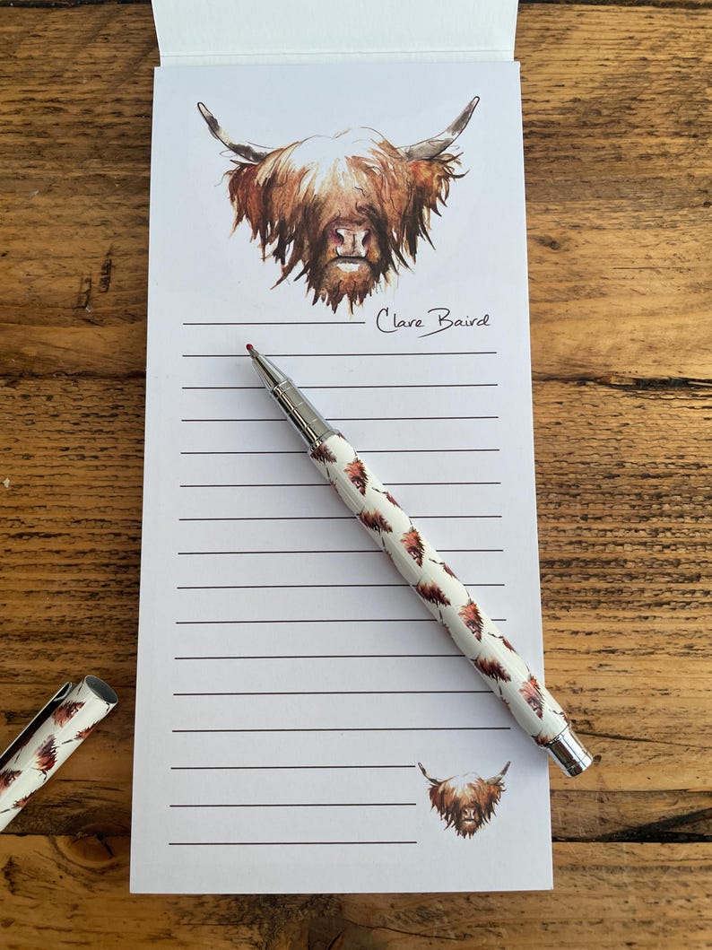Pu&ograve; includere: Un blocco note bianco con un'illustrazione di mucca Highland marrone e bianca in alto. Il blocco note ha carta rigata e una penna argentata con un motivo di mucca Highland marrone e bianca. Il blocco note &egrave; firmato "Clare Baird".