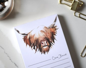 Puede incluir: Un bloc de notas blanco con una ilustración de acuarela de una vaca Highland marrón y blanca con pelo largo. El bloc de notas tiene una sección rayada en la parte inferior con el texto "Clare Baird" escrito en tinta negra.