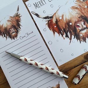 Pu&ograve; includere: Un blocco note bianco con carta rigata e un'illustrazione di una mucca delle Highlands marrone e bianca. Il blocco note ha una firma a mano che dice "Clare Baird". Una penna con un motivo di mucca delle Highlands marrone e bianca &egrave; appoggiata sul blocco note. Un biglietto bianco con un'illustrazione di una mucca delle Highlands marrone e bianca e il testo "Merry Christmas" &egrave; sullo sfondo.