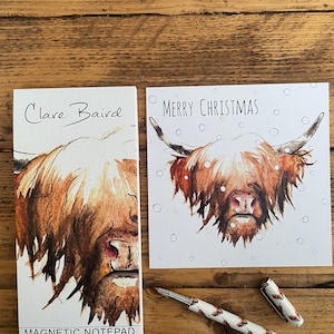 Pu&ograve; includere: Un biglietto bianco con il testo "Merry Christmas" e un'illustrazione ad acquerello di una mucca delle Highlands con manto marrone e macchie bianche. La mucca guarda lo spettatore con un'espressione seria. Il biglietto &egrave; circondato da punti bianchi che assomigliano a fiocchi di neve. Il biglietto &egrave; accanto a un blocco note magnetico bianco con il testo "Clare Baird" e un'illustrazione ad acquerello della testa di una mucca delle Highlands. Il blocco note ha il testo "Magnetic Notepad" in basso. Una penna bianca con un motivo marrone e bianco &egrave; appoggiata su una superficie di legno.