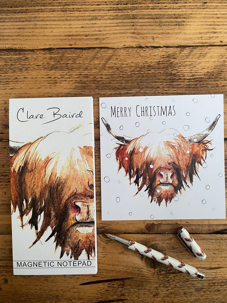 Pu&ograve; includere: Un biglietto bianco con il testo "Merry Christmas" e un'illustrazione ad acquerello di una mucca delle Highlands con manto marrone e macchie bianche. La mucca guarda lo spettatore con un'espressione seria. Il biglietto &egrave; circondato da punti bianchi che assomigliano a fiocchi di neve. Il biglietto &egrave; accanto a un blocco note magnetico bianco con il testo "Clare Baird" e un'illustrazione ad acquerello della testa di una mucca delle Highlands. Il blocco note ha il testo "Magnetic Notepad" in basso. Una penna bianca con un motivo marrone e bianco &egrave; appoggiata su una superficie di legno.