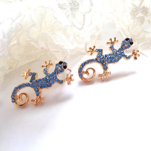 Peut inclure: Une paire de boucles d'oreilles en forme de lézard dorées. Les lézards sont ornés de strass bleus et d'yeux noirs. Les boucles d'oreilles ont un fermoir à tige. Elles sont posées sur une surface blanche.
