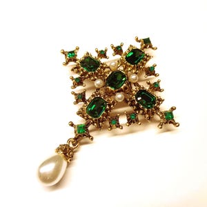Può includere: Una spilla color oro a forma di croce con molteplici pietre verde smeraldo a taglio rettangolare e quadrato, piccole perle bianche rotonde e un singolo grande ciondolo di perla bianca a goccia appeso alla base dell'oggetto.