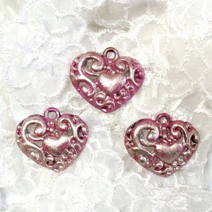 Könnte beinhalten: Drei verzierte herzförmige Anhänger mit einem Used-Look-Finish in Rosa und Silber. Jeder Anhänger hat ein kleineres Herz in der Mitte und dekorative Schnörkel. Die Anhänger haben oben eine Schlaufe zum Basteln.