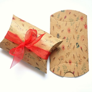 Puede incluir: Dos cajas de almohada de papel kraft con un estampado floral. Una caja está atada con una cinta roja. Las cajas tienen un diseño botánico con flores y hojas en tonos de rosa, verde y marrón.