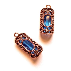 Può includere: Due ciondoli rettangolari color rame con una pietra centrale blu a forma ovale. I ciondoli hanno un design a filigrana attorno alla pietra blu e un piccolo anello nella parte superiore per appenderli. Lo sfondo è bianco.