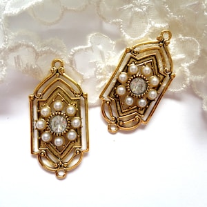 Peut inclure: Deux pendentifs en métal doré ornés. Chaque pendentif présente un strass clair central entouré de petites perles de fausse couleur blanche. Les pendentifs ont un design Art Déco avec des formes géométriques et une boucle en haut pour suspendre.