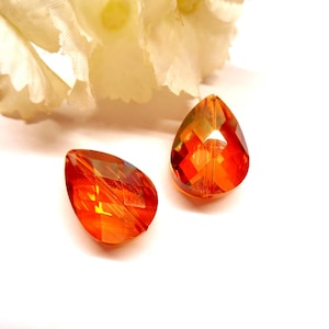 Pode incluir: Duas contas laranja em forma de gota, facetadas e com efeito degradê, refletindo a luz. As contas estão sobre um fundo branco, com um elemento floral branco e macio no canto superior esquerdo. Adequado para a criação de joias.