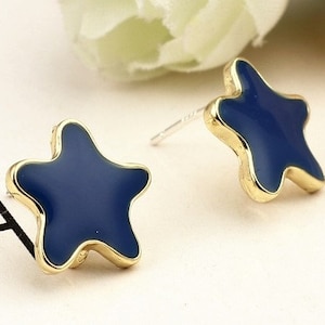 Puede incluir: Un par de pendientes de botón con forma de estrella. Los pendientes presentan un acabado esmaltado azul marino con un borde dorado. Los pendientes están sobre un fondo suave y neutro.