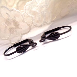 Peut inclure: Deux apprêts de boucles d'oreilles à levier noirs. Chaque apprêt comporte un fil incurvé et un charm décoratif en forme de goutte. Les apprêts sont posés sur un fond blanc avec un détail de dentelle dans le coin supérieur gauche.