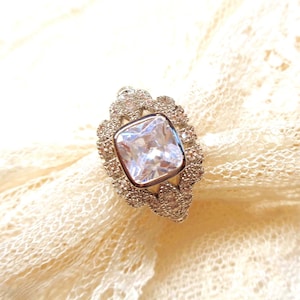 Puede incluir: Un anillo plateado con una gran piedra preciosa clara y cuadrada en el centro. La piedra preciosa está rodeada por un halo de piedras claras más pequeñas. El anillo tiene un diseño ornamentado y detallado alrededor de la piedra principal y la banda.
