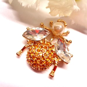 Peut inclure: Broche dorée en forme d'abeille, ornée d'une tête de perle et d'ailes en cristal transparent. Le corps est recouvert de petits cristaux orange-jaune. La broche a des accents rouges sur les pattes, sur fond blanc.