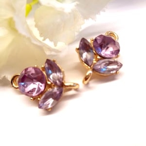 Peut inclure: Une paire de boucles d'oreilles dorées avec un motif floral. Chaque boucle d'oreille présente une grosse pierre précieuse rose, ronde et facettée, entourée de petites pierres précieuses violettes en forme de marquise. Les boucles d'oreilles sont posées sur un fond blanc avec une fleur blanche floue.
