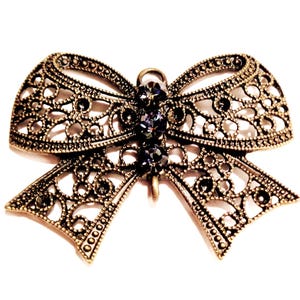 Op de afbeelding: Een gedetailleerde, antieke strikbroche in bronskleur. De strik heeft een open ontwerp met ingewikkelde patronen en is versierd met verschillende kleine, donkerpaarse edelstenen in het midden. De broche staat tegen een witte achtergrond.