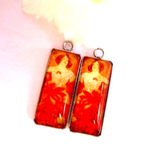 Peut inclure: Deux pendentifs rectangulaires avec un design art nouveau. Chaque pendentif représente une femme en robe fluide avec des accents floraux rouges. Les pendentifs ont un cadre en métal argenté et une petite boucle en haut pour suspendre.