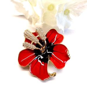 Könnte beinhalten: Eine blumenförmige Brosche mit leuchtend roten Emaille-Blütenblättern und schwarzen Akzenten in der Mitte. Das Stück ist mit goldfarbenen Metallrändern und einer Ansammlung klarer, funkelnder Strasssteine als Staubgefäß verziert.