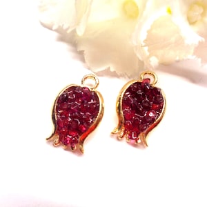 Pode incluir: Dois pingentes em forma de romã com uma moldura de metal dourado e uma incrustação cristalina vermelho-escura texturizada. Cada pingente inclui um pequeno laço circular no topo para fixação, exibindo uma silhueta detalhada de fruta.