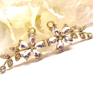 Op de afbeelding: Twee goudkleurige bloemen oorbellen met heldere kristallen accenten. Elke oorbel heeft een bloemdesign met meerdere druppelvormige kristallen en een delicate blad tak met kleinere kristallen. De oorbellen zijn geplaatst tegen een zachte, witte achtergrond.
