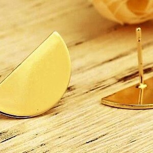 Può includere: Due orecchini color oro, ciascuno a forma di semicerchio. Uno è un pezzo pieno, mentre l'altro ha un perno e un piccolo anello. Gli orecchini sono esposti su una superficie di legno chiaro.