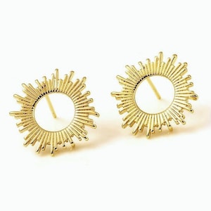 Peut inclure: Une paire de boucles d'oreilles dorées. Chaque boucle d'oreille présente un motif de soleil avec un cercle central ouvert et des pointes rayonnantes. Les boucles d'oreilles sont posées sur un fond blanc, mettant en valeur leurs détails.