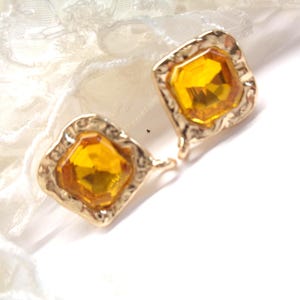 Peut inclure: Une paire de boucles d'oreilles carrées dorées avec une bordure texturée. Chaque boucle d'oreille présente une grosse pierre précieuse facettée de couleur ambre. Les boucles d'oreilles sont posées sur un fond blanc avec de la dentelle délicate.