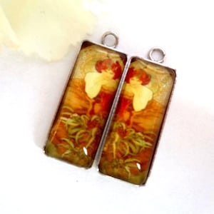 Peut inclure: Deux pendentifs rectangulaires avec un design art nouveau vintage. Chaque pendentif représente une femme aux cheveux roux et un châle crème, sur un fond doré et chaleureux. Les pendentifs ont des cadres en métal argenté et une boucle en haut pour les suspendre.