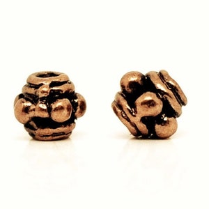Peut inclure: Deux perles en métal de couleur bronze. L'une est en forme de tonneau avec une série de rainures horizontales et de protubérances arrondies. L'autre perle est un amas de formes arrondies. Les deux perles sont sur fond blanc.
