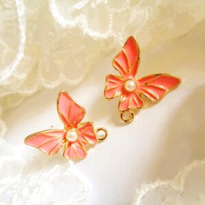Peut inclure: Deux breloques en forme de papillon avec des bords en métal doré et des ailes en émail rose corail. Chaque breloque présente une petite perle blanche artificielle ronde au centre et un anneau métallique circulaire en bas pour la fixation.