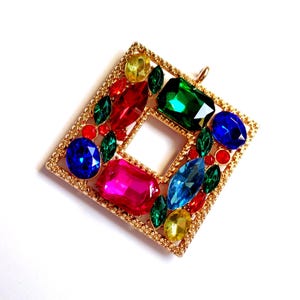 Könnte beinhalten: Ein goldfarbener Anhänger in quadratischer Form mit einem zentralen offenen Quadrat. Der Anhänger ist mit bunten Edelsteinen in Rot, Grün, Blau, Gelb und Pink besetzt. Die Edelsteine sind in einem dekorativen Goldrahmen gefasst.