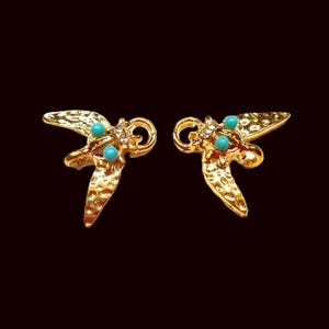 Peut inclure: Une paire de boucles d'oreilles dorées avec une finition texturée. Chaque boucle d'oreille présente un motif d'oiseau stylisé avec des accents turquoise et de petites pierres transparentes. Les boucles d'oreilles sont posées sur un fond marron foncé.
