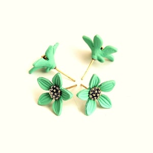 Pode incluir: Quatro brincos de pressão em forma de flor. Os brincos são de cor verde-água clara com um centro dourado escuro e pinos dourados. Estão dispostos sobre um fundo branco.
