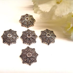 Puede incluir: Cinco casquillos para cuentas de metal plateado con un diseño floral circular calado. Cada pieza presenta un orificio central y recortes simétricos intrincados en forma de pétalos que irradian hacia afuera, creando una apariencia texturizada.