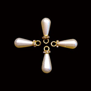 Peut inclure: Quatre breloques en forme de goutte d'eau nacrées, avec des accents dorés et de petits strass transparents. Chaque breloque a une boucle en haut pour l'attacher à des bijoux. Les breloques sont disposées en forme de croix sur fond noir.