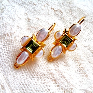 May include: A pair of gold-toned earrings featuring a central square green gemstone, surrounded by four white, oval-shaped pearls. The earrings have a hook closure and are set against a white lace background.